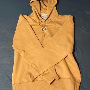 Beige hoodie från Tommy Hilfiger - Säljer en snygg beige hoodie från Tommy Hilfiger med broderad logga på bröstet. Tröjan har huva med dragsko och långa ärmar. Perfekt för en avslappnad stil. Skulle säga den sitter som L 