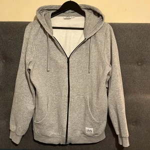 Ljusgrå hoodie med dragkedja från And1 - Säljer en ljusgrå hoodie från And1 med dragkedja framtill och två stora fickor. Tröjan har huva med dragsko och ribbade muddar vid ärmslut och nederkant. Perfekt för en avslappnad stil.