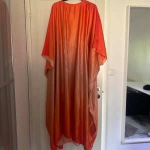 Silkesklänning i orange ombre - Fjäril-klänning i silkesmaterial med ombre-effekt. Klänningen har vid passform och är ärmlös med rund halsringning. Det finns ett resår band på insidan som man lägger bakom ryggen för att få den mer figursydd. Prislapp kvar.