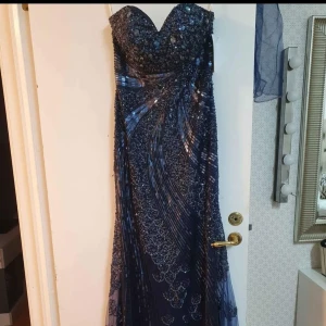 Marinblå långklänning med paljetter - Säljer en marinblå långklänning med glittriga paljetter och pärldetaljer. Klänningen har sweetheart-urringning och är axelbandslös, perfekt för bal eller fest. Materialet är glansigt och skimrande, vilket ger en lyxig känsla.