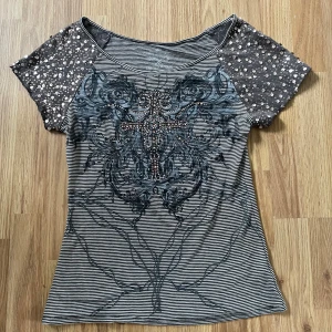 Randig topp med paljettärmar och mönster - Säljer en svartvit randig topp med korta ärmar täckta av paljetter. Framsidan har ett stort dekorativt mönster i grått och svarta toner samt glittriga detaljer. Toppen har en feminin passform och är perfekt för dig som gillar unika detaljer.