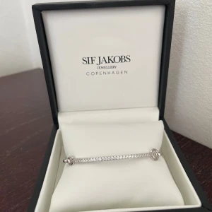 Silverfärgat armband från Sif Jakobs - SIF JACOBS CAPIZZI BANGLE  Armband i rhodinerat 925 Sterling silver med polerad yta och handplacerade facettslipade vita zirkoner. Armbandets mått: Inre diameter 47x58 mm, bredd 2.6 mm, tjocklek 2.3 mm