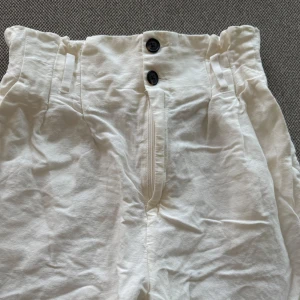 Vita byxor med hög midja - Säljer ett par vita byxor med hög midja och papperspåse-linning. Byxorna har två knappar framtill och diskreta fickor i sidorna. De är perfekta för varma dagar. Skärp saknas. Storlek 36
