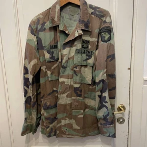 Camouflage militärjacka U.S. Army - Säljer en klassisk militärjacka i camouflage från U.S. Army med broderade märken och namnlapp. Jackan har lång ärm, flera fickor framtill och patch med AIRBORNE på axeln. Perfekt för dig som gillar army-stil.