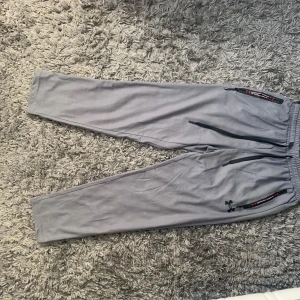 Grå träningsbyxor från Under Armour - Snygga grå träningsbyxor från Under Armour med svarta dragkedjor vid fickorna och elastisk midja. Perfekta för gymmet eller löprundan. Byxorna har en smalare passform och är tillverkade i ett mjukt syntetmaterial.