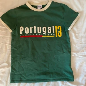 Grön Portugal t-shirt från Bershka - Grön t-shirt från Bershka med vit kontrastkrage och ärmslut. Tryck på bröstet med texten 'Portugal 13' och årtalet 1998, samt stor siffra 13 på ryggen. Perfekt för dig som gillar sportig stil.