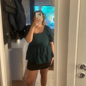 Mörkgrön blus med puffärm och öppen rygg - Säljer en mörkgrön blus med kort puffärm och volang nedtill. Toppen har öppen rygg med två knytband och rundad halsringning. 