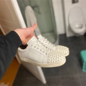 Loubs - Säljer ett par vita sneakers från Christian Louboutin med coola nitar framtill och ikonisk röd sula. Skorna har snörning och en klassisk rund tå. Perfekta för dig som vill sticka ut med din stil. Kan även byta skor 