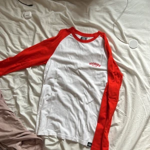 Vit och röd långärmad tröja från Dickies - Säljer en långärmad tröja från Dickies i storlek medium. Tröjan är vit med röda ärmar och röd krage, klassisk baseballstil. Tryck med Dickies-logga och text på bröstet. Perfekt för en avslappnad och sportig look.