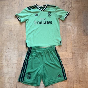 Real Madrid fotbollströja och shorts - Säljer en grön Real Madrid fotbollströja från Adidas i storlek M. Tröjan har svarta detaljer, korta ärmar och klassisk v-ringad krage med svarta och vita ränder. Materialet är lätt och andas, perfekt för träning. Matchande gröna shorts med svarta ränder ingår.