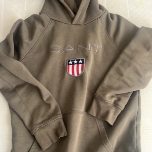 Olivgrön hoodie från GANT med brodyr - Olivgrön hoodie från GANT med broderad logga på bröstet. Hoodien har luva, känguruficka och ribbade muddar. Mjuk insida och relaxed fit, perfekt för chill dagar. Snygg detalj med amerikanskt inspirerat emblem.