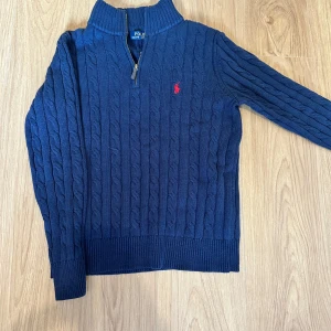Marinblå kabelstickad tröja Polo Ralph Lauren - Kabelstickad marinblå tröja från Polo Ralph Lauren med halv dragkedja och röd broderad logga på bröstet. Tröjan har hög krage och långa ärmar, perfekt för kyliga dagar. Klassisk och stilren design som funkar till det mesta. Den är M men passar som S