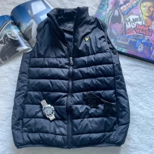 Svart dunväst från Lyle & Scott - Svart quiltad dunväst från Lyle & Scott med gul broderad logga på bröstet. Västen har hög krage och dragkedja framtill.