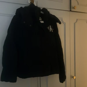 Svart pufferjacka Calvin Klein Jeans - Svart pufferjacka från Calvin Klein Jeans med stor CK-logga på bröstet. Jackan har huva, dragkedja och tryckknappar framtill. Jackan har fickor med knappar storlek M