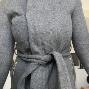 Grå omlottkappa med bälte och dragkedja - Snygg grå kappa i omlottmodell med bälte i midjan och coola dragkedjor i silver. Jackan har hög krage och långa ärmar, perfekt för kyliga dagar. Materialet känns mjukt och stilrent, och detaljerna ger en modern vibe.