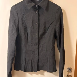 Svart skjorta från SHEIN med struktur - Stilren svart skjorta från SHEIN med lång ärm och markerad krage. Skjortan har en strukturerad yta och knäppning framtill med svarta knappar. Figursydd modell som ger en snygg siluett, perfekt till jeans eller kjol.