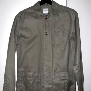 Olivgrön overshirt från John Henric - Snygg olivgrön overshirt från John Henric med klassisk krage, tre stora fickor framtill och knäppning med bruna knappar. Tillverkad i bomull, perfekt för lager-på-lager och en clean look. Passar dig som gillar stilrena och enkla plagg.