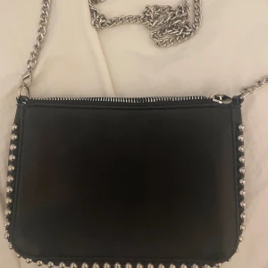 Svart axelväska med kedja  - Snygg svart axelväska i skinnimitation med silverfärgade pärlor längs kanterna. Går att ta bort kedjan och använda som ex necessär. 