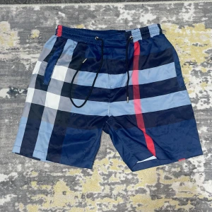 Blårutiga Burberry badshorts - Snygga Burberry badshorts med klassiskt rutmönster i olika nyanser av blått, vitt och en röd detalj. Shortsen har elastisk midja med snörning och praktiska fickor. Perfekta för sommaren och strandhäng, tillverkade i lätt polyester.