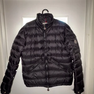 Svart Moncler Agay dunjacka med dragkedja - Säljer en svart dunjacka från Moncler med hög krage och snygga detaljer. Jackan har två snedställda fickor med dragkedja och Moncler-logga på ärmen. Materialet är glansigt och quiltat, perfekt för kalla dagar. Stängs med dragkedja framtill. Storlek : 1/S men kan passa XS