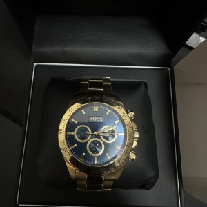 Guldig BOSS klocka med blå urtavla - Stilren klocka från Hugo Boss med guldig länk och blå urtavla. Klockan har tre mindre urtavlor, tydliga index och kronograf-funktion. Snygg kombination av guld och blått som ger en lyxig känsla. Levereras i originalask med BOSS-logga. Nypris 6500. Finns några små repor som inte syns om man inte kollar noggrant. Vattenresistant klocka