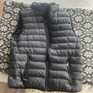 Svart dunväst från Polo Ralph Lauren - Säljer en svart dunväst från Polo Ralph Lauren med diskret broderad logga på bröstet. Västen har två dragkedjefickor framtill, hög krage och quiltad design. Perfekt att slänga över en hoodie eller tröja när det är kyligt ute. Man får med en Ralph lauren påse 