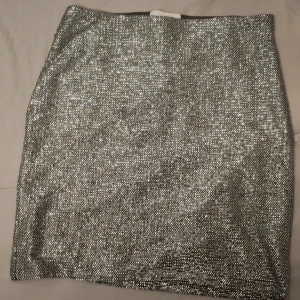 Glittrig silverkjol från H&M - Snygg kort glittrig kjol från H&M i silver. Kjolen har en rak passform och är täckt av små glittrande paljetter som verkligen fångar ljuset. Perfekt för dig som vill sticka ut och glänsa lite extra.