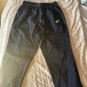 Svarta träningsbyxor från Asics - Aldrig använt. Svarta träningsbyxor från Asics med elastisk midja och diskret logga på låret. Byxorna är tillverkade i ett lätt och syntetiskt material som passar perfekt för sport och rörelse. Klassisk rak passform med fickor på sidorna.