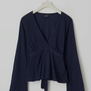 Mörkblå omlottopp med v-ringning - Supersnygg mörkblå omlottopp från Gina Tricot med v-ringning och långa ärmar. Toppen har knyt i midjan och är tillverkad i ett lätt, strukturerat tyg. Perfekt för dig som gillar stilrena och trendiga plagg.