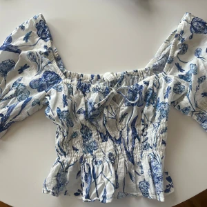Blommig offshoulder blus från H&M - Supersöt blommig blus från H&M i vitt och blått. Offshoulder-modell med puffiga korta ärmar, smockad midja och volangkant nertill. Blusen har knytning framtill och är gjord i ett lätt bomullstyg, perfekt för varma dagar.