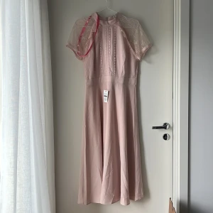 Ljusrosa spetsklänning från Liquorish - Superfin ljusrosa klänning från Liquorish med spetsärmar och dekorativa spetsdetaljer framtill. Klänningen har hög hals, dragkedja i ryggen och är knälång med lätt utsvängd kjol. Perfekt för dig som gillar romantisk och feminin stil.