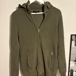 Olivgrön stickad hoodie från Indicode - Säljer en olivgrön stickad hoodie från Indicode som är extremt snygg och eftertraktad!, med dragkedja framtill och två fickor med dragkedja. Tröjan har en quiltad huva i blankt material och långa ärmar. Perfekt för dig som gillar chill och stilrena plagg 