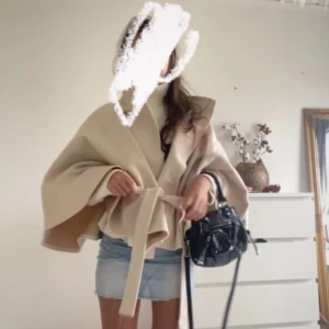 Beige poncho kappa med bälte - Säljer en beige poncho med breda ärmar och snygg omlottknäppning med bälte i midjan. Den är ifrån Nelly, men slutsåld och svår att få tag på. 