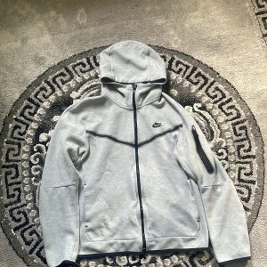 Nike tech fleece  - Snygg ljusgrå hoodie från Nike med hel dragkedja och svart logga på bröstet. Tröjan har huva, svarta detaljer och en praktisk ficka med dragkedja på ärmen. Perfekt för chill eller träning, riktigt clean och sportig look.