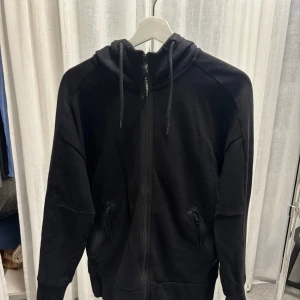 Svart hoodie med goggles från C.P. Company - Svart hoodie från C.P. Company med hel dragkedja, två fickor med dragkedja och ikoniska goggles på huvan. Tröjan har snörning i huvan och är långärmad. 