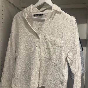 Vit broderad skjorta från Zara - Superfin vit skjorta från Zara i bomull med broderade blommor och diskret struktur. Skjortan har lång ärm, bröstficka och avslappnad passform. Perfekt för en fräsch och somrig look.