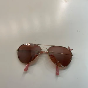 Guldiga aviator-solglasögon med fjäril - Snygga aviator-solglasögon med guldigt metallbåge och rosatonade glas. Skalmarna är rosa och på ena linsen sitter en liten rosa fjärilsdetalj som ger extra vibe. Perfekta för att lyfta din stil i sommar.
