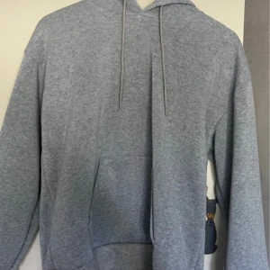 Grå hoodie med huva och ficka - En klassisk grå hoodie med huva och snörning. Tröjan har en stor magficka och långa ärmar. Perfekt för chill dagar eller när du vill ha en enkel och stilren look.