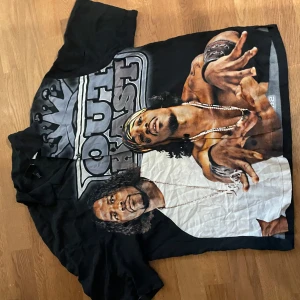 Outkast svart skjorta med tryck - Svart skjorta med stort färgstarkt Outkast-tryck på framsidan och albumlista på baksidan. Motivet visar båda medlemmarna i Outkast, detaljer som halskedja och bandana. T-shirten har rund hals och korta ärmar, tillverkad i mjuk bomull.