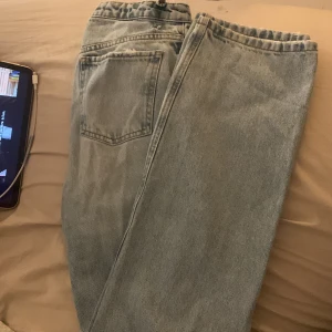 Ljusblå ripped jeans från NA-KD - Säljer ett par ljusblå jeans från NA-KD i storlek 34. Jeansen har en snygg ripped detalj på ena benet, klassisk femficksmodell och raka ben. Perfekta för en chill och trendig look.