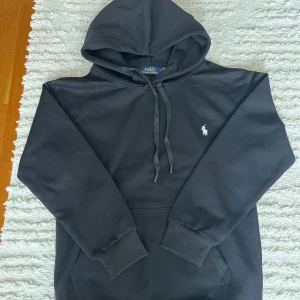 Svart Ralph Lauren Hoodie – Storlek S - Klassisk hoodie från Polo Ralph Lauren i svart färg med den ikoniska vita polo-loggan broderad på bröstet. 	•	Storlek: S, men passformen är lite rymlig och kan även passa om du vanligtvis bär M beroende på önskad fit. 	•	Material: Mjuk och bekväm bomullsblandning. 	•	Fickor: Känguruficka framtill. 	•	Huva med justerbara snören.  Ett tidlöst plagg som passar både till vardags och mer avslappnade tillfällen.