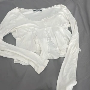 Vit långärmad blus med spets från Gina Tricot - Supersöt vit långärmad blus från Gina Tricot med volang nertill, spetsdetaljer längs kanterna och en liten rosett framtill. Blusen har en feminin och romantisk look med rundad halsringning och croppad passform. Perfekt för dig som gillar mjuka och gulliga vibes.