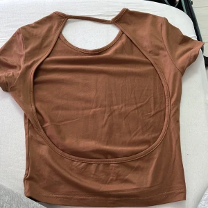 Brun t-shirt med öppen rygg - Snygg brun t-shirt med korta ärmar och en stor, rund öppning i ryggen som ger en cool och unik look. Materialet känns mjukt och stretchigt, perfekt för dig som gillar att sticka ut. Passformen är normal och färgen är varm brun.
