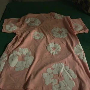 Rosa blommig t-shirt Denim Tears M - Säljer en rosa t-shirt från Denim Tears i storlek M. T-shirten har vita blommiga tryck över hela plagget och är gjord i mjuk bomull. Modellen är klassisk med korta ärmar och rund hals. Perfekt för dig som gillar streetwear med unik design.