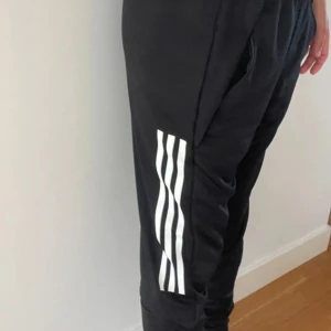 Svarta Adidas träningsbyxor S - Svarta träningsbyxor från Adidas med vita ränder längs benen och Adidas-logga på låret. Byxorna har elastisk midja, smal passform och är gjorda i mjukt polyester-material. Perfekta för gymmet eller chill hemma. Snygg sportig look med fickor på sidorna.