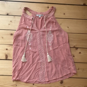 Rosa broderad linnetopp - Superfin ljusrosa linnetopp från Papaya med broderade mönster framtill och dekorativa snören med tofsar. Toppen är ärmlös och har en avslappnad passform, perfekt för varma dagar. Snygga detaljer längs halsringningen och ett boho-inspirerat uttryck. Hör av er vid minsta fråga💞😇