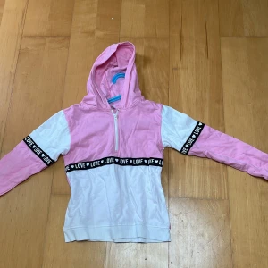 Rosa och vit hoodie med LOVE-band - Snygg hoodie i rosa och vitt med svart band där det står LOVE runt armar och midja. Tröjan har huva, lång ärm och en half zip-dragkedja framtill. Perfekt för dig som gillar en sportig och trendig stil.