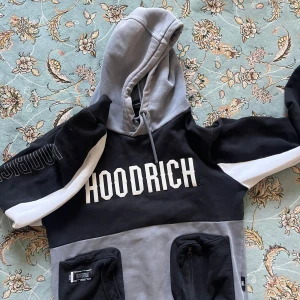 Hoodrich hoodie svart/grå M - Säljer en snygg hoodie från Hoodrich i storlek medium. Tröjan har svart och grå färgblock, vit logga framtill och coola detaljer på ärmen. Utrustad med huva och snörning samt två fickor med dragkedja. Perfekt för streetwear-stilen.