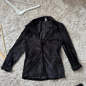 Svart omlottkavaj från & Other Stories - Snygg svart kavaj från & Other Stories Paris Atelier med omlott och bälte i midjan. Gjord i glansig sammet med subtilt ribbad struktur, klassisk krage och långa ärmar. Perfekt för dig som gillar stilrena och trendiga plagg med lite extra lyxig känsla.