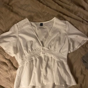 Vit blus med spetsdetaljer  - Säljer en söt vit blus med v-ringning, korta ärmar och fina spetsdetaljer längs axlar och midja. Blusen har knappar framtill och en lätt volang nedtill som ger en romantisk vibe. Perfekt att styla med jeans eller kjol! Denna är i storlek L men passar mig bra som har storlek s - m vanligtvis